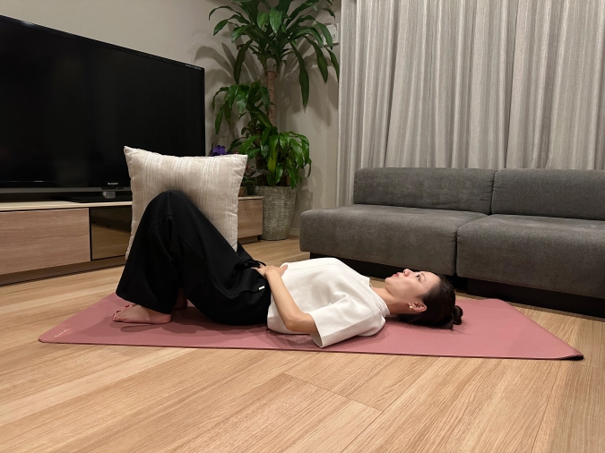 仰向けになって両脚を立てる。脚は骨盤幅に開き、両膝の間にクッションを挟む。