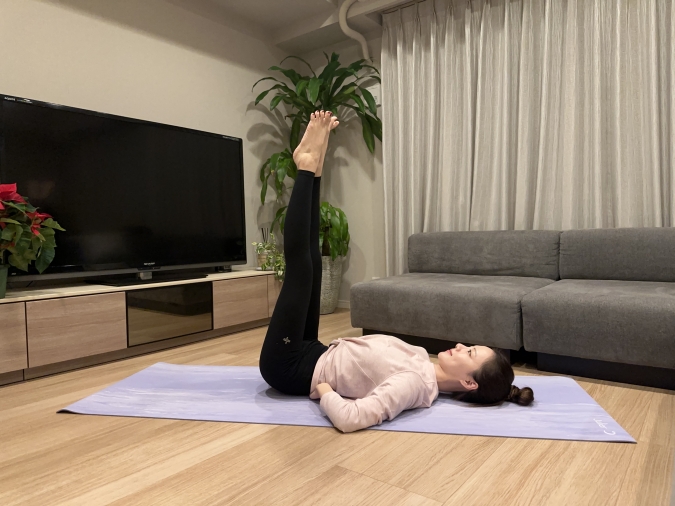 仰向けになり、両脚を天井方向に向かって伸ばす。つま先を少し外に開き、かかと同士を軽く寄せてVの字を作る。両手はお尻と腰の間に入れ、下腹を引き締める。