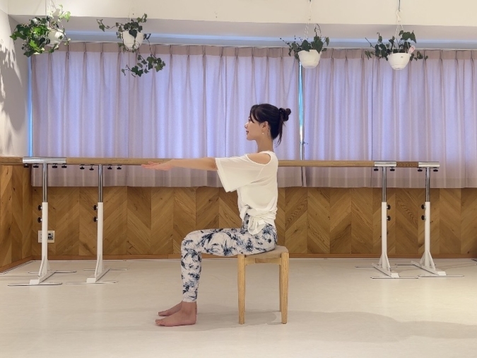 椅子に浅く座り、両手を前へ伸ばし、息を吸いながら背筋を伸ばす。